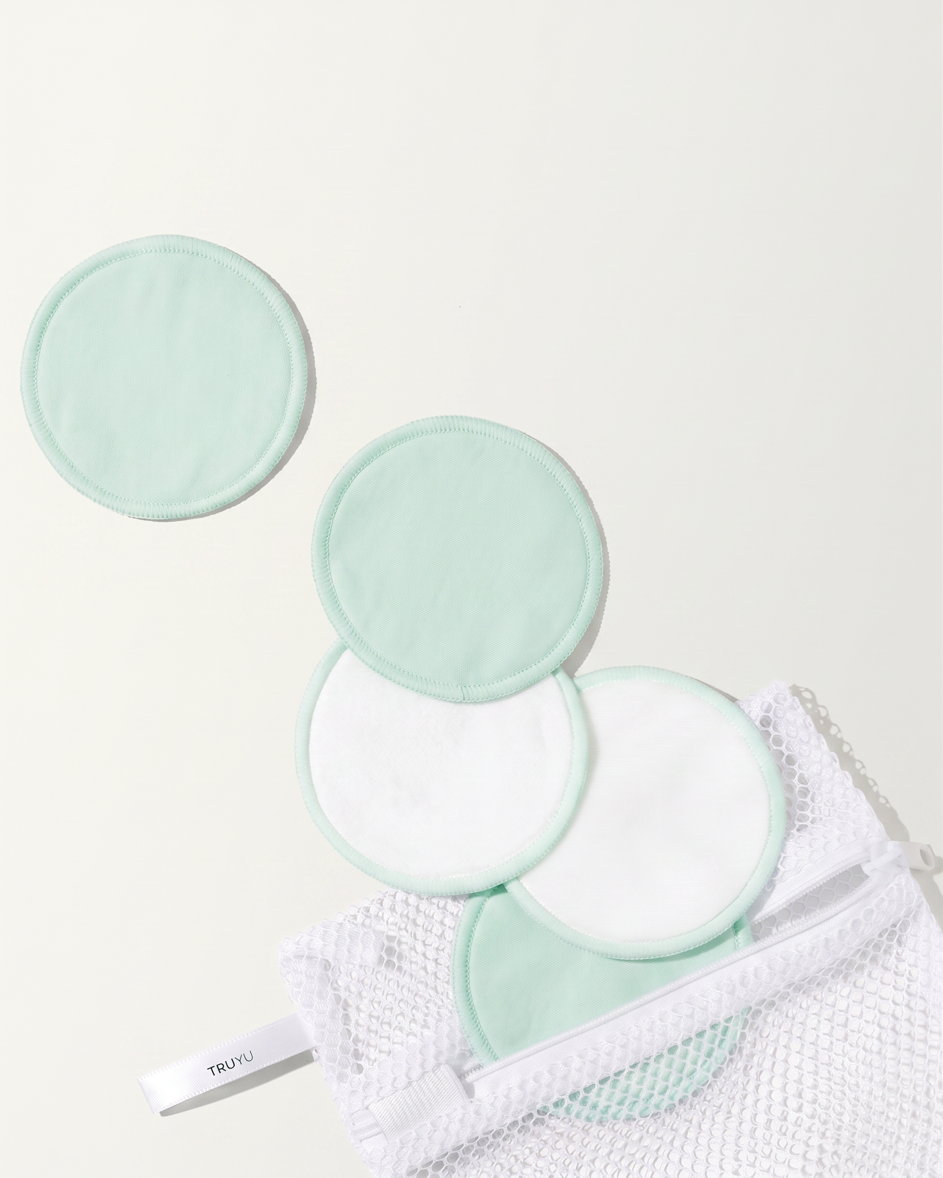 Set of five light green and white reusable makeup remover pads with a white mesh bag on a light cream background