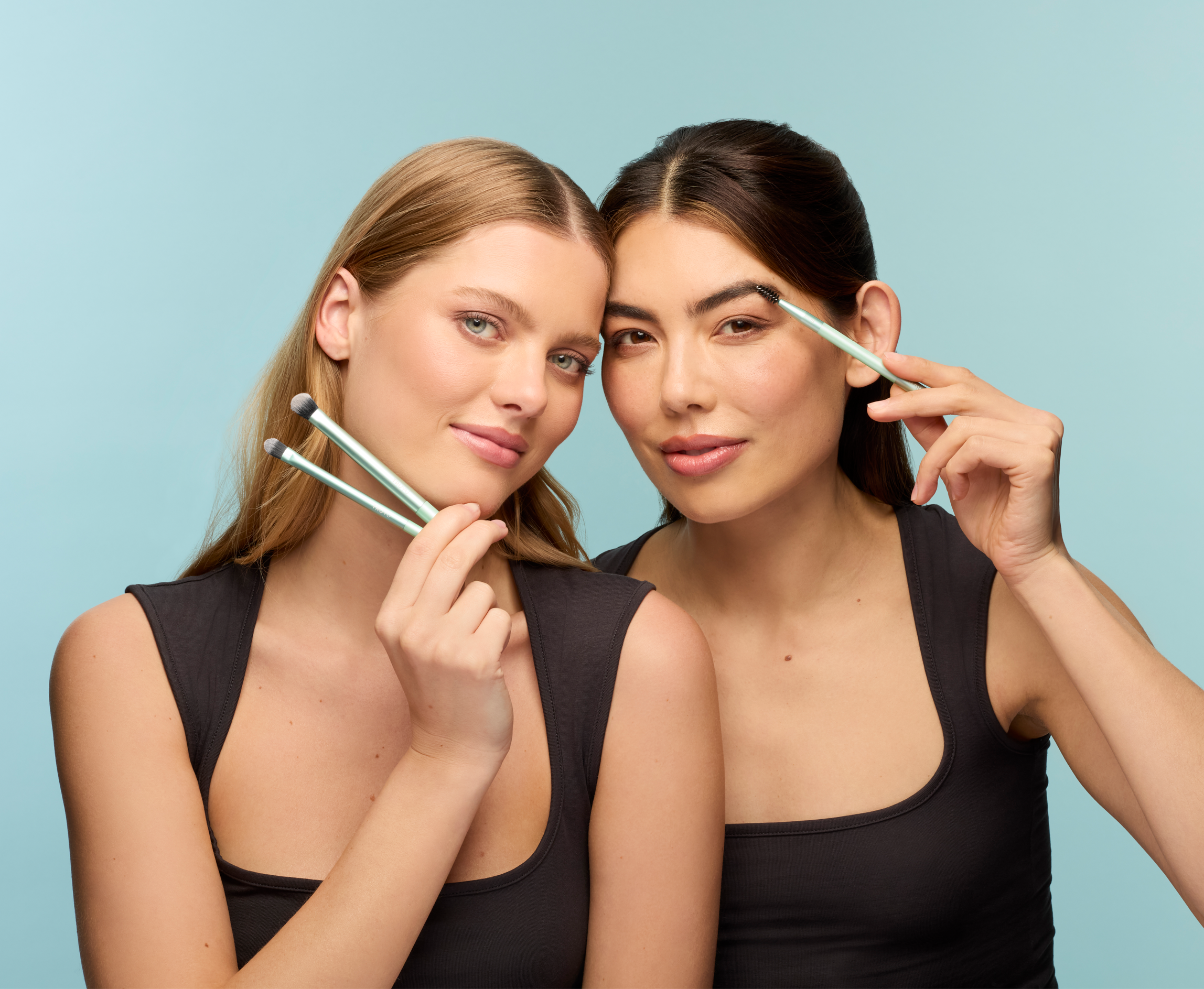 2 Models in black tops, holding TRUYU Brow Brush and Eyeshadow Brushes near face. Light blue background. 