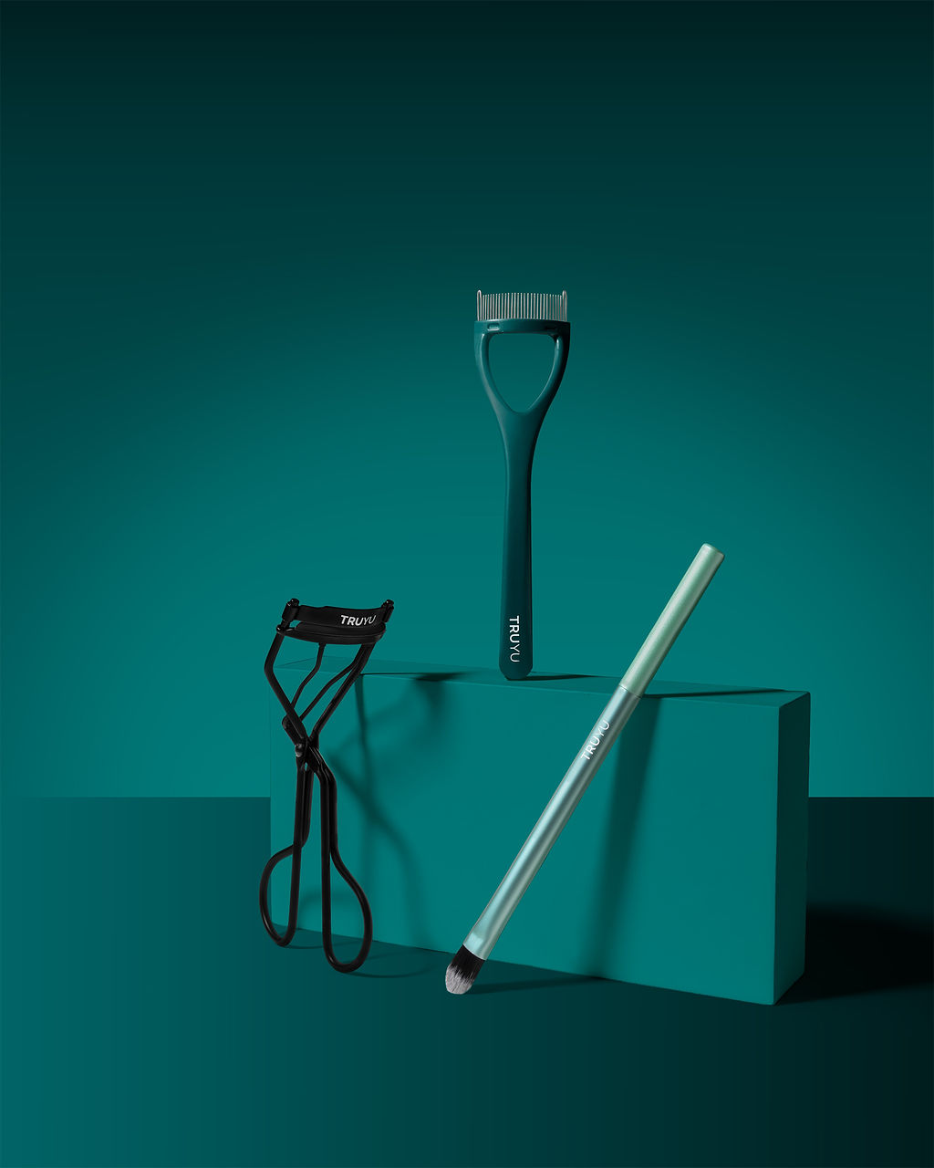 TRUYU Complete Eye Set out of pack. Including an eyeshadow brush, eyelash curler, and eyelash comb on a teal background.