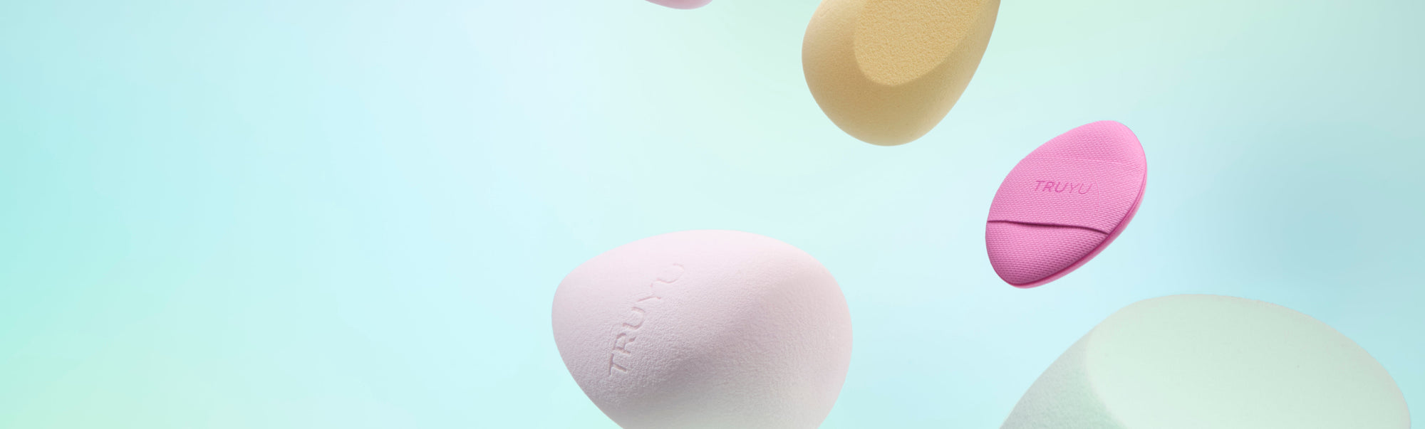Six pastel-colored silicone beauty sponges floating at different heights against a soft mint green and light blue gradient background. The sponges are in shades of pink, pale yellow, light blue, and mint green, with the brand name 'TRULU' subtly embossed on some of them.
