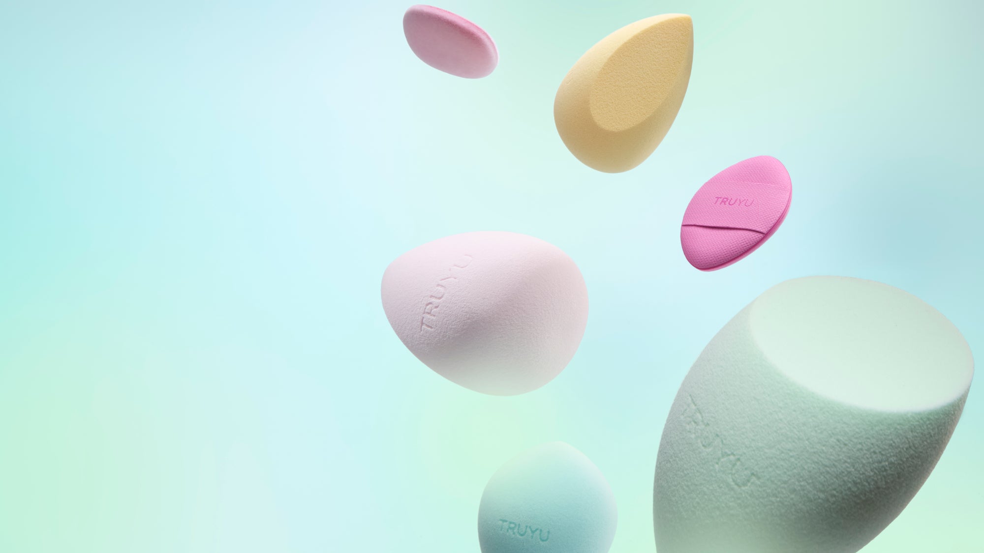 Six pastel-colored silicone beauty sponges floating at different heights against a soft mint green and light blue gradient background. The sponges are in shades of pink, pale yellow, light blue, and mint green, with the brand name 'TRULU' subtly embossed on some of them.