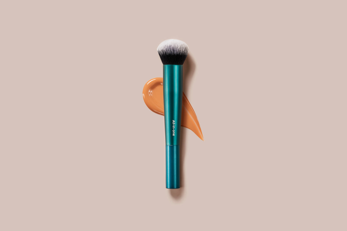 A makeup brush with a metallic teal handle and white bristles, positioned vertically over a peach-colored cosmetic swatch on a solid beige background.